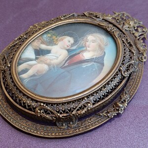 K&ouml;nnte beinhalten: Antikes, oval gerahmtes Kunstwerk mit religi&ouml;ser Szene. Das Bild zeigt eine Frau, die ein Kind h&auml;lt, mit einem Engel im Hintergrund. Der Rahmen ist verziert, mit Bronze-Finish und aufwendigen Details, ca. 15 cm.