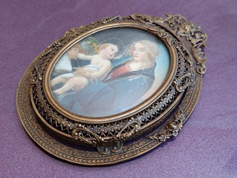 K&ouml;nnte beinhalten: Antikes, oval gerahmtes Kunstwerk mit religi&ouml;ser Szene. Das Bild zeigt eine Frau, die ein Kind h&auml;lt, mit einem Engel im Hintergrund. Der Rahmen ist verziert, mit Bronze-Finish und aufwendigen Details, ca. 15 cm.