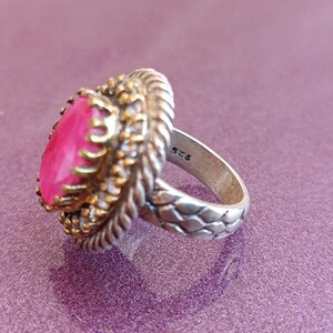 Op de afbeelding: Een zilveren ring met een roze edelsteen gezet in een filigraan ontwerp van goudkleur. De band is getextureerd en heeft het nummer "925" gegraveerd aan de binnenkant.