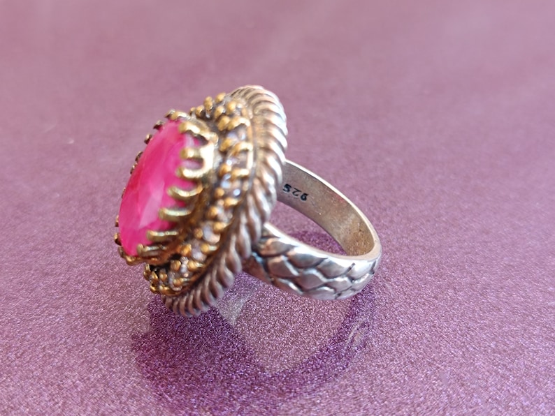 Op de afbeelding: Een zilveren ring met een roze edelsteen gezet in een filigraan ontwerp van goudkleur. De band is getextureerd en heeft het nummer "925" gegraveerd aan de binnenkant.