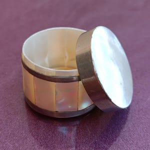 Op de afbeelding: Een klein, rond, wit parelmoer doosje met een zilveren rand en een scharnierend deksel. Het doosje is open en de binnenkant is zichtbaar.