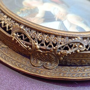 K&ouml;nnte beinhalten: Ein antiker ovaler Bilderrahmen mit einem detaillierten, verzierten goldfarbenen Metallrand. Der Rahmen zeigt ein kleines Gem&auml;lde mit einem verschwommenen Bild einer Person. Der Rahmen hat einen dekorativen schmetterlingsf&ouml;rmigen Akzent.