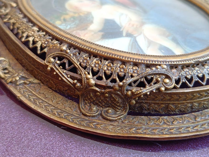 K&ouml;nnte beinhalten: Ein antiker ovaler Bilderrahmen mit einem detaillierten, verzierten goldfarbenen Metallrand. Der Rahmen zeigt ein kleines Gem&auml;lde mit einem verschwommenen Bild einer Person. Der Rahmen hat einen dekorativen schmetterlingsf&ouml;rmigen Akzent.