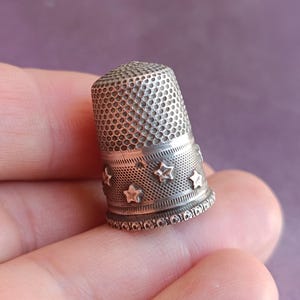 May include: A silver thimble with a perforated top and a band of star-shaped cutouts. The thimble is held in a hand, showcasing its detailed design and craftsmanship. This sewing tool is a classic item for any sewing kit.