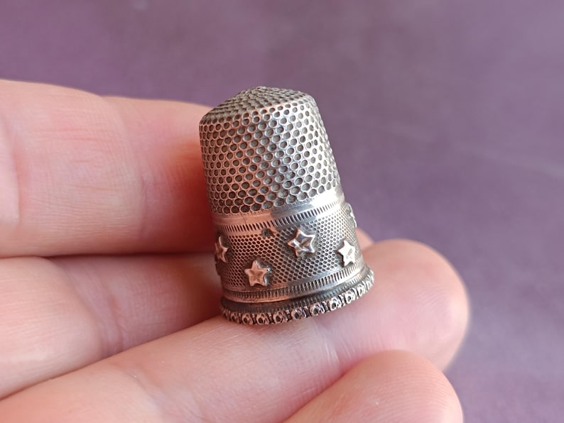 May include: A silver thimble with a perforated top and a band of star-shaped cutouts. The thimble is held in a hand, showcasing its detailed design and craftsmanship. This sewing tool is a classic item for any sewing kit.