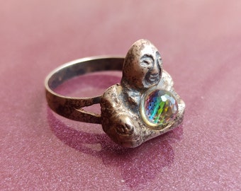 May include: Silver ring featuring a detailed sculpture of a seated Buddha holding a round iridescent gemstone.