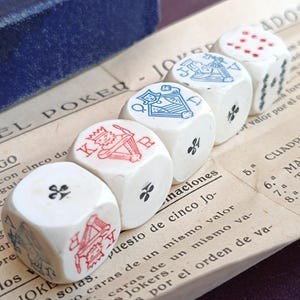 May include: Five white poker dice with red and blue suit symbols and numbers, arranged on a vintage paper with Spanish text. The dice show King, Queen, and Jack symbols, along with other markings. A blue box is in the background.
