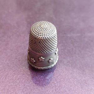 May include: A silver thimble with a textured top and a band of star-shaped cutouts around the base. The thimble is set against a purple background, showcasing its intricate design and craftsmanship. A sewing accessory.