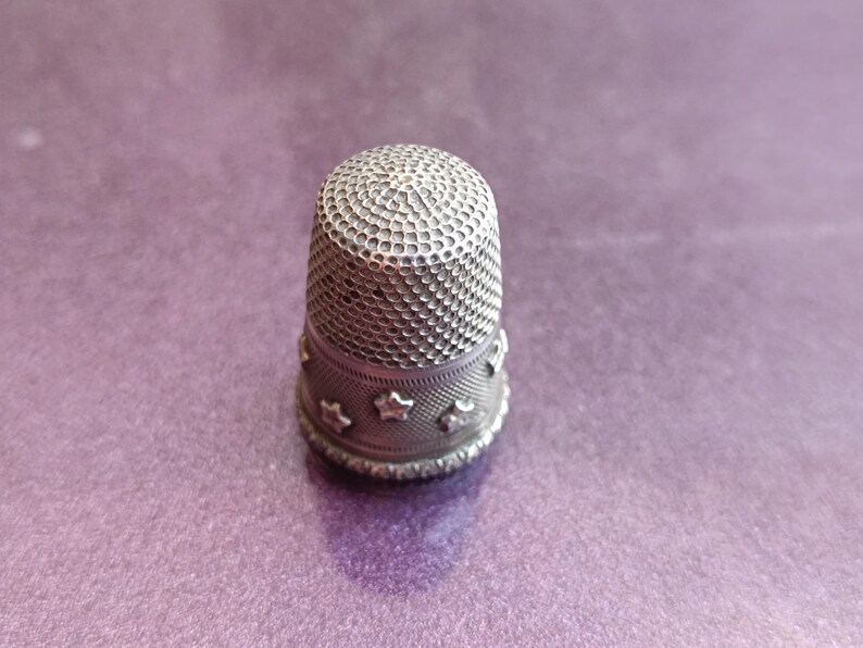 May include: A silver thimble with a textured top and a band of star-shaped cutouts around the base. The thimble is set against a purple background, showcasing its intricate design and craftsmanship. A sewing accessory.