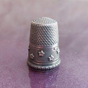 May include: A silver thimble with a textured surface and a band of star-shaped embellishments. The thimble is a sewing tool, designed to protect the finger while pushing a needle through fabric. The thimble is approximately 1 inch tall.