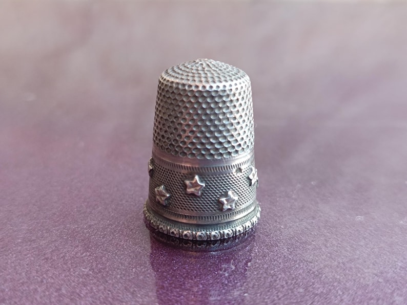 May include: A silver thimble with a textured surface and a band of star-shaped embellishments. The thimble is a sewing tool, designed to protect the finger while pushing a needle through fabric. The thimble is approximately 1 inch tall.