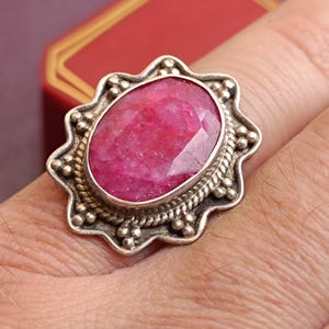 Op de afbeelding: Een zilveren ring met een grote, ovaal geslepen robijn. De robijn heeft een levendige roze-rode kleur en is gezet in een decoratieve zilveren bezel met een touwachtige rand en kraalaccenten. De ring zit om een vinger.