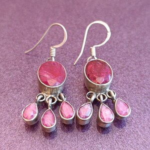 May include: A pair of silver dangle earrings with pink gemstone drops. The earrings feature a round pink gemstone with a silver setting and three smaller pink gemstone drops hanging from the bottom.