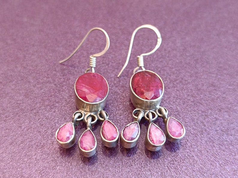 May include: A pair of silver dangle earrings with pink gemstone drops. The earrings feature a round pink gemstone with a silver setting and three smaller pink gemstone drops hanging from the bottom.