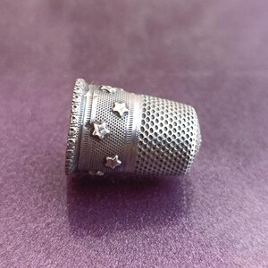 May include: A silver thimble with a decorative band of stars and a perforated body. The thimble is designed for sewing and crafting, offering protection for the fingertip. The thimble is made of metal and is set against a purple background.