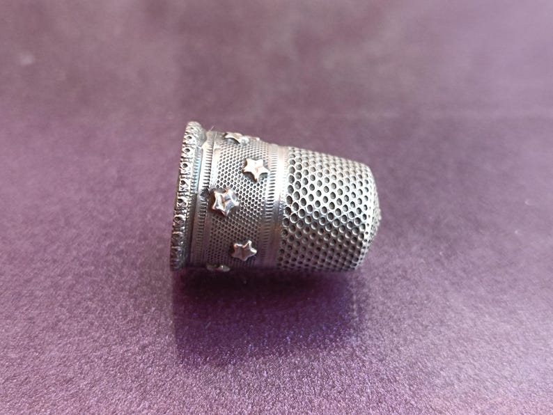May include: A silver thimble with a decorative band of stars and a perforated body. The thimble is designed for sewing and crafting, offering protection for the fingertip. The thimble is made of metal and is set against a purple background.