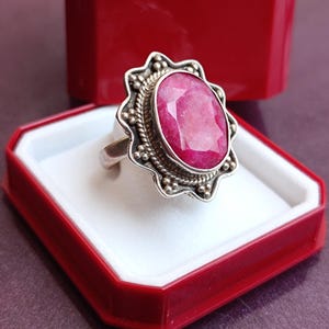 Op de afbeelding: Een zilveren ring met een grote, ovaal geslepen robijn edelsteen. De robijn heeft een levendige roze-rode kleur en is gezet in een decoratieve bezel. De ring wordt gepresenteerd in een rode sieradendoos met een wit interieur.