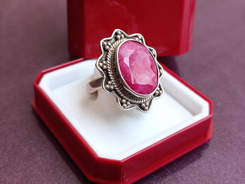 Op de afbeelding: Een zilveren ring met een grote, ovaal geslepen robijn edelsteen. De robijn heeft een levendige roze-rode kleur en is gezet in een decoratieve bezel. De ring wordt gepresenteerd in een rode sieradendoos met een wit interieur.