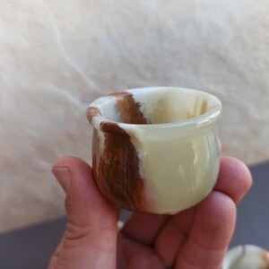 K&ouml;nnte beinhalten: Eine kleine, wei&szlig;e und braune Onyx-Schale mit abgerundeter Form. Die Schale ist aus Naturstein gefertigt und hat eine glatte, polierte Oberfl&auml;che.