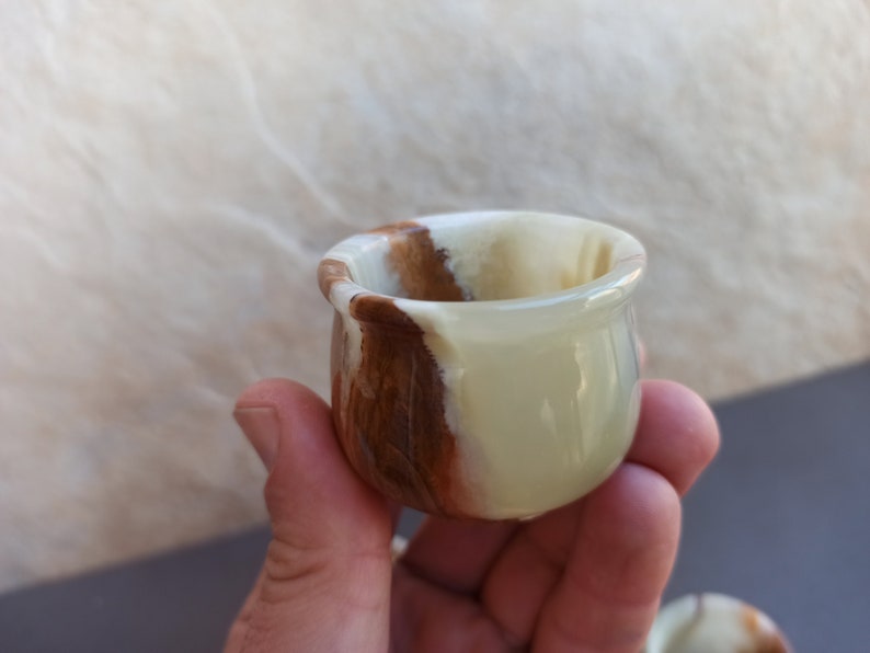 K&ouml;nnte beinhalten: Eine kleine, wei&szlig;e und braune Onyx-Schale mit abgerundeter Form. Die Schale ist aus Naturstein gefertigt und hat eine glatte, polierte Oberfl&auml;che.