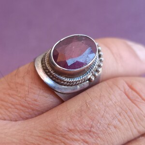 Pode incluir: Um anel de prata com uma grande pedra preciosa oval, facetada, de cor p&uacute;rpura, engastada em uma moldura de prata. O anel tem uma banda decorativa com um design de corda e pequenas contas.