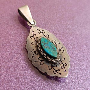 May include: A silver pendant with a turquoise stone center. The pendant has a leaf-like shape with intricate cut-out designs and a braided rope border around the stone. The bail is a simple rectangular shape.