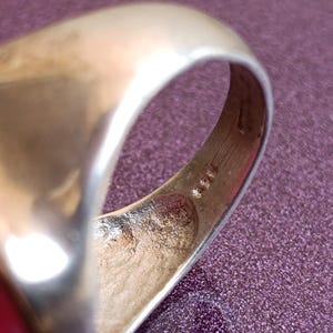Op de afbeelding: Close-up van een goudkleurige ring. De ring heeft een gladde, afgeronde band en een platte bovenkant. De binnenkant van de band toont markeringen. De ring is geplaatst tegen een paarse achtergrond.