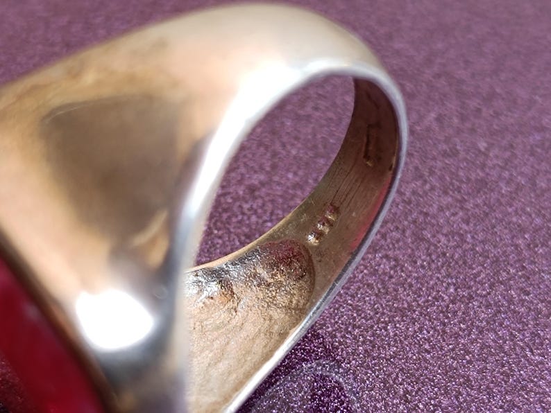Op de afbeelding: Close-up van een goudkleurige ring. De ring heeft een gladde, afgeronde band en een platte bovenkant. De binnenkant van de band toont markeringen. De ring is geplaatst tegen een paarse achtergrond.