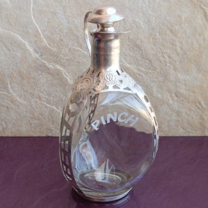 May include: A clear glass decanter with a silver-plated top and a silver-plated cage around the body. The decanter has the word "PINCH" etched on the side.
