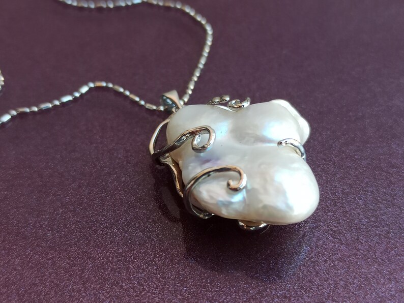 May include: A silver chain necklace with a white baroque pearl pendant. The pearl is set in a silver cage with delicate swirls.