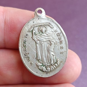 May include: An oval silver pendant with a detailed relief of a robed figure holding an object, inscribed with "S. VICENTE FERRER". The pendant has a small loop at the top for hanging. The pendant is likely made of silver.