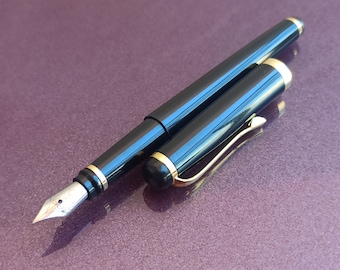 May include: A black fountain pen with gold accents and a fine nib. The pen is closed and lying on a purple surface.