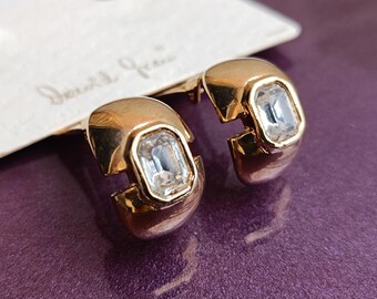 Op de afbeelding: Een paar goudkleurige oorbellen met een heldere, rechthoekige edelsteen in het midden. De oorbellen hebben een uniek, open ontwerp. De achtergrond is dieppaars en een label met de naam "David Yurman" is zichtbaar.
