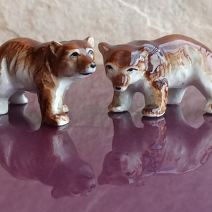 Puede incluir: Dos figuritas de cerámica de osos, una marrón y blanca, la otra blanca con marcas marrones. Los osos se miran, con la cabeza ligeramente inclinada. Están de pie sobre una superficie morada.