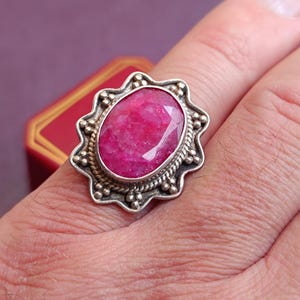 Op de afbeelding: Een zilveren ring met een grote, ovaal geslepen robijn. De robijn heeft een levendige rozerode kleur en is gezet in een sierlijke zilveren zetting. De ring zit om een vinger.