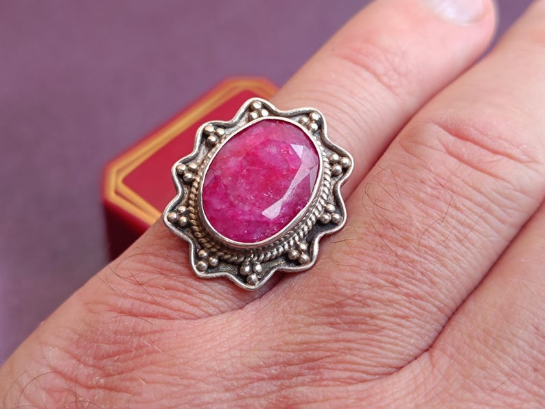 Op de afbeelding: Een zilveren ring met een grote, ovaal geslepen robijn. De robijn heeft een levendige rozerode kleur en is gezet in een sierlijke zilveren zetting. De ring zit om een vinger.