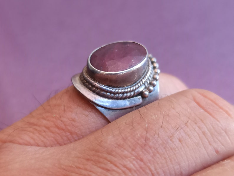 Pode incluir: Um anel de prata com uma grande pedra preciosa ovalada de cor p&uacute;rpura engastada no centro. O anel tem detalhes intrincados de prata em torno da pedra preciosa.