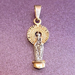 Könnte beinhalten: Ein goldfarbener Anhänger mit einer detaillierten Figur in einem Sonnenstrahl-Design. Der Anhänger hat eine kleine Schlaufe für eine Kette und ist vor einem lila Hintergrund platziert. Die Figur scheint ein Baby zu halten.