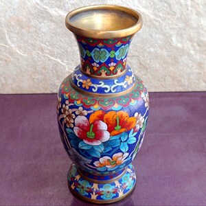 Op de afbeelding: Een cloisonné vaas met een levendige blauwe achtergrond en ingewikkelde bloemmotieven in rood, oranje, wit en groen. De vaas heeft een messing rand aan de bovenkant en een messing voet, met gedetailleerd metaaldraadwerk rondom elk geëmailleerd deel.