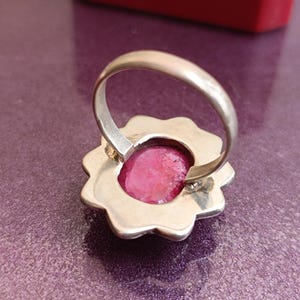 Op de afbeelding: Een zilveren ring met een bloemvormige zetting. De ring heeft een grote, ovaalvormige, felroze edelsteen. De ring is geplaatst op een paarse ondergrond, met een rood object zichtbaar op de achtergrond.