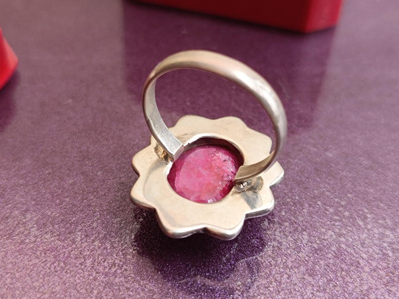 Op de afbeelding: Een zilveren ring met een bloemvormige zetting. De ring heeft een grote, ovaalvormige, felroze edelsteen. De ring is geplaatst op een paarse ondergrond, met een rood object zichtbaar op de achtergrond.