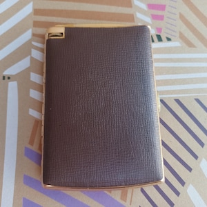 May include: A brown leather cigarette case with a gold trim. The case has a textured surface and a hinged lid.