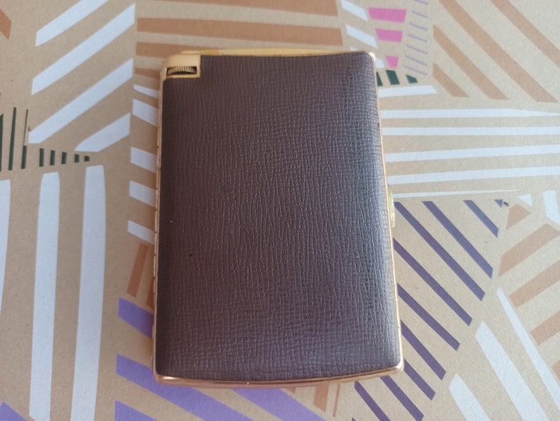 May include: A brown leather cigarette case with a gold trim. The case has a textured surface and a hinged lid.