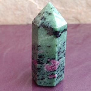 May include: A green and black ruby zoisite crystal point. The crystal has a hexagonal base and a pointed top. The green color is speckled with black and pink inclusions. The crystal is a natural stone.