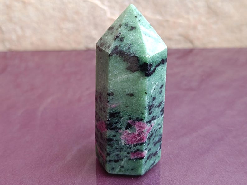 May include: A green and black ruby zoisite crystal point. The crystal has a hexagonal base and a pointed top. The green color is speckled with black and pink inclusions. The crystal is a natural stone.
