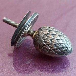 May include: Silver bottle stopper featuring a detailed pine cone design. The stopper has a threaded base and a decorative top.