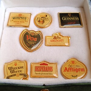 May include: A collection of eight gold-colored beer bottle cap pins. The pins feature various beer brands, including Murphy's Irish Red, Fischer, Guinness, Vos, Adelscott, Wieckse Witte, Desperados, and Affligem.