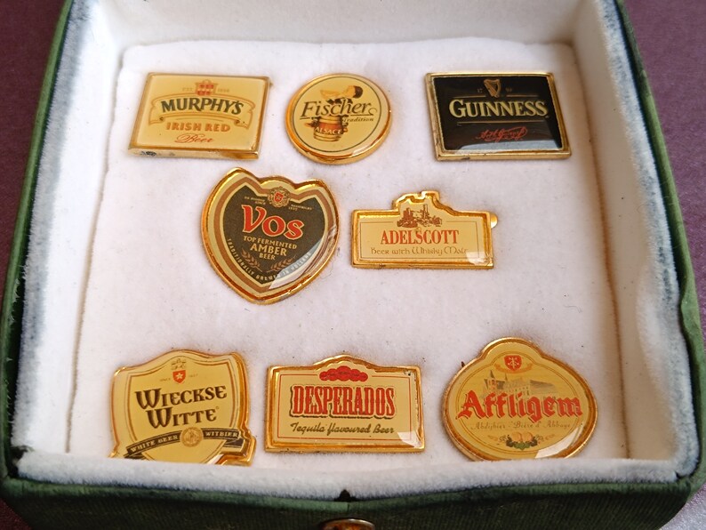 May include: A collection of eight gold-colored beer bottle cap pins. The pins feature various beer brands, including Murphy's Irish Red, Fischer, Guinness, Vos, Adelscott, Wieckse Witte, Desperados, and Affligem.