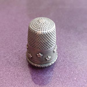 May include: A silver thimble with a textured, dimpled top and a band of star-shaped cutouts around the base. The thimble is set against a purple background, showcasing its detailed craftsmanship. A sewing tool.