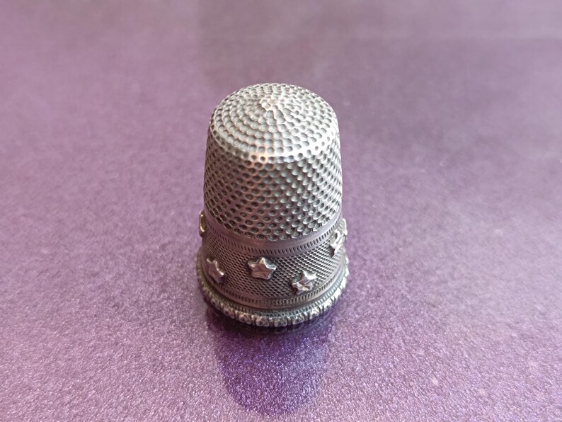 May include: A silver thimble with a textured, dimpled top and a band of star-shaped cutouts around the base. The thimble is set against a purple background, showcasing its detailed craftsmanship. A sewing tool.
